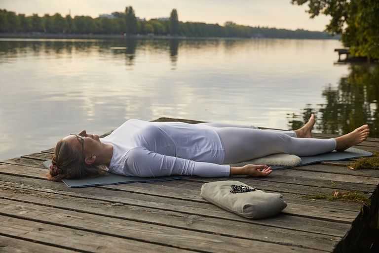Frau in schlichter weißer Kleidung ausgestreckt in Shavasana auf hölzernem Steg im Hintergrund See und Bäume, Kundalini Yoga Meditation und Entspannung in Hannover Maschsee